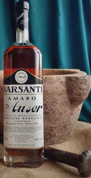 Bottiglia di Amaro Auser Barsanti accanto a un grande mortaio in pietra con pestello, su tessuto grezzo e sfondo verde petrolio.