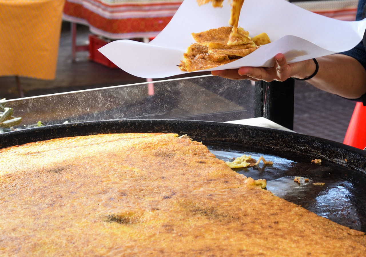 Una persona serve una porzione di cecina appena cotta, tagliata a pezzi e disposta su un foglio di carta, accanto alla grande teglia usata per la cottura.
