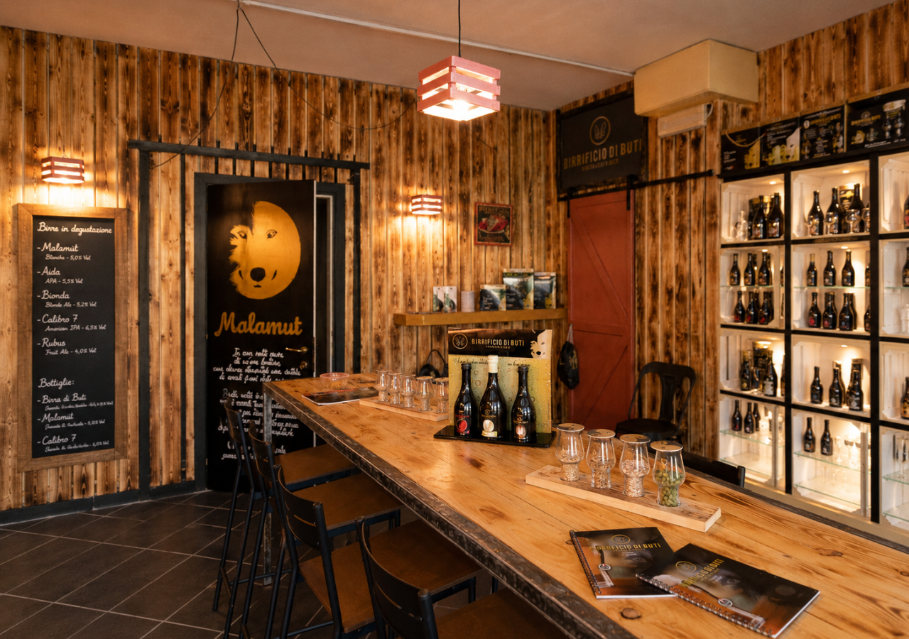 Interno del Birrificio di Buti con pareti in legno, lungo tavolo per le degustazioni, bottiglie di birra artigianale, bicchieri e scaffali espositivi illuminati.