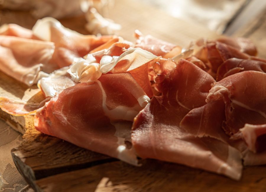 Tagliere di legno con sottili fette di prosciutto crudo e altri salumi, illuminati dalla luce calda del sole su una tavola apparecchiata.