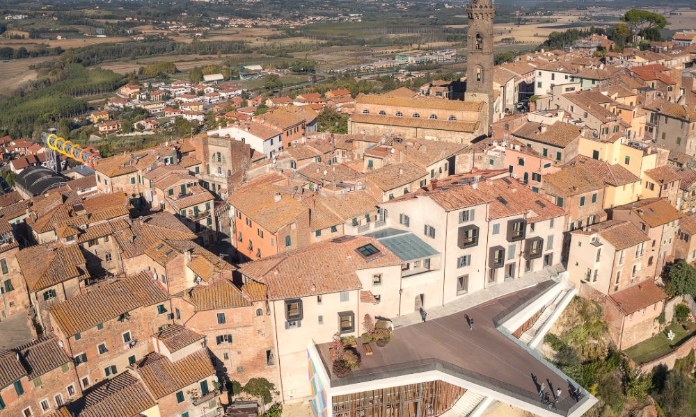 Veduta aerea del borgo di Peccioli, nelle Terre di Pisa, con tetti in laterizio, campanile storico, terrazza panoramica e paesaggio collinare sullo sfondo.