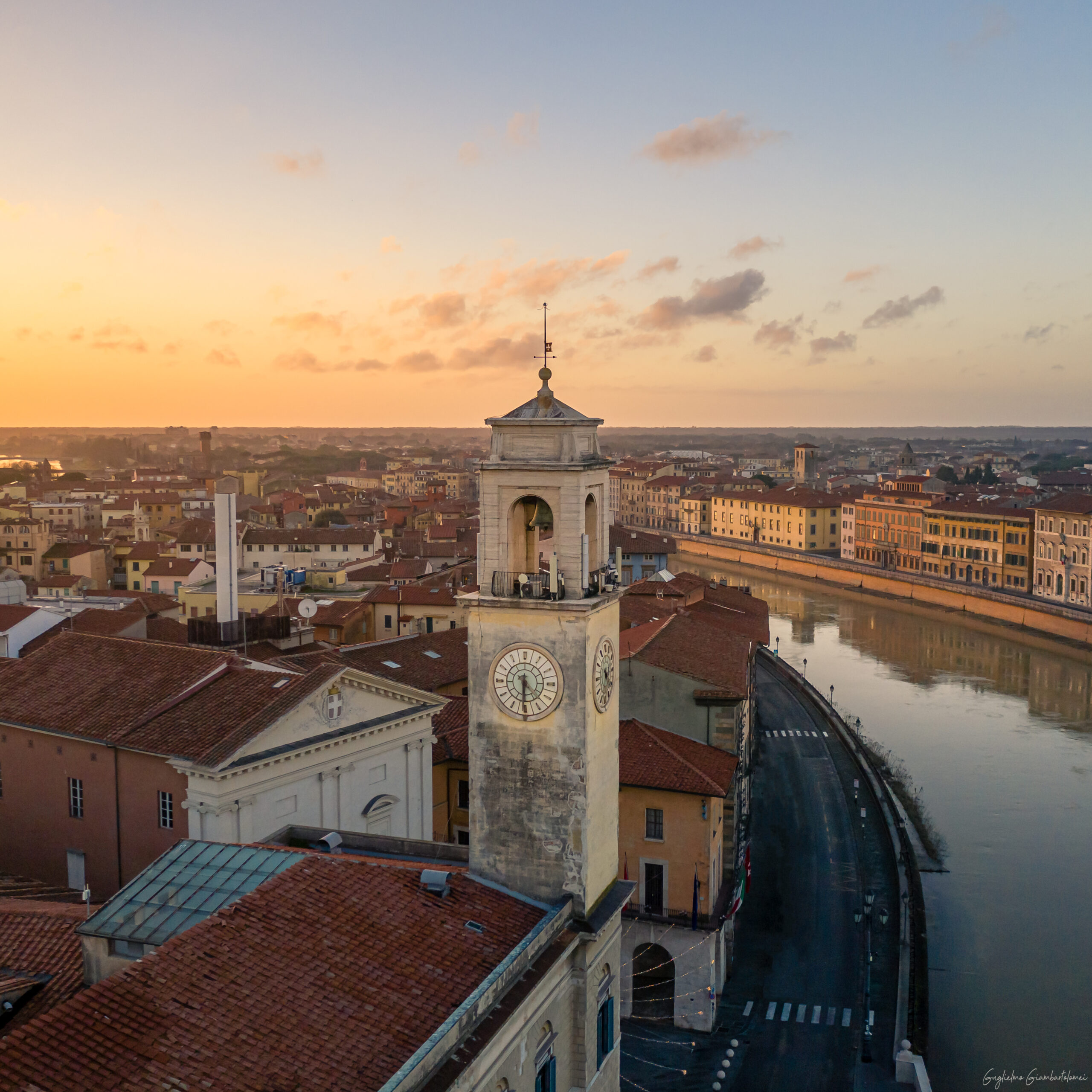 Veduta aerea di Pisa al tramonto, con la torre dell’orologio di Palazzo Pretorio in primo piano e i Lungarni lungo il fiume Arno sullo sfondo.
