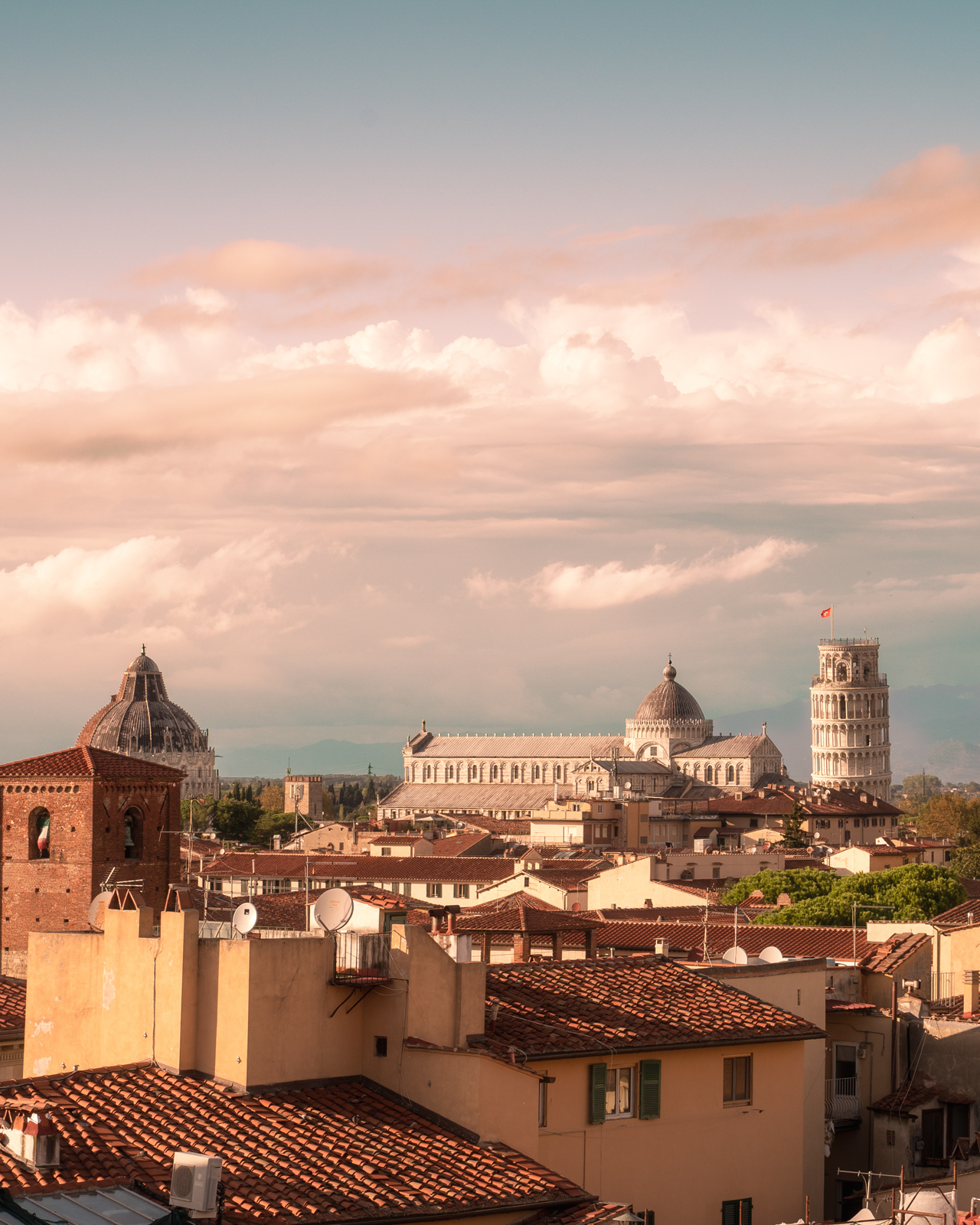 Veduta di Pisa dai tetti, con Piazza dei Miracoli sullo sfondo, la Torre pendente, il Duomo e le cupole illuminate dalla luce del tramonto.
