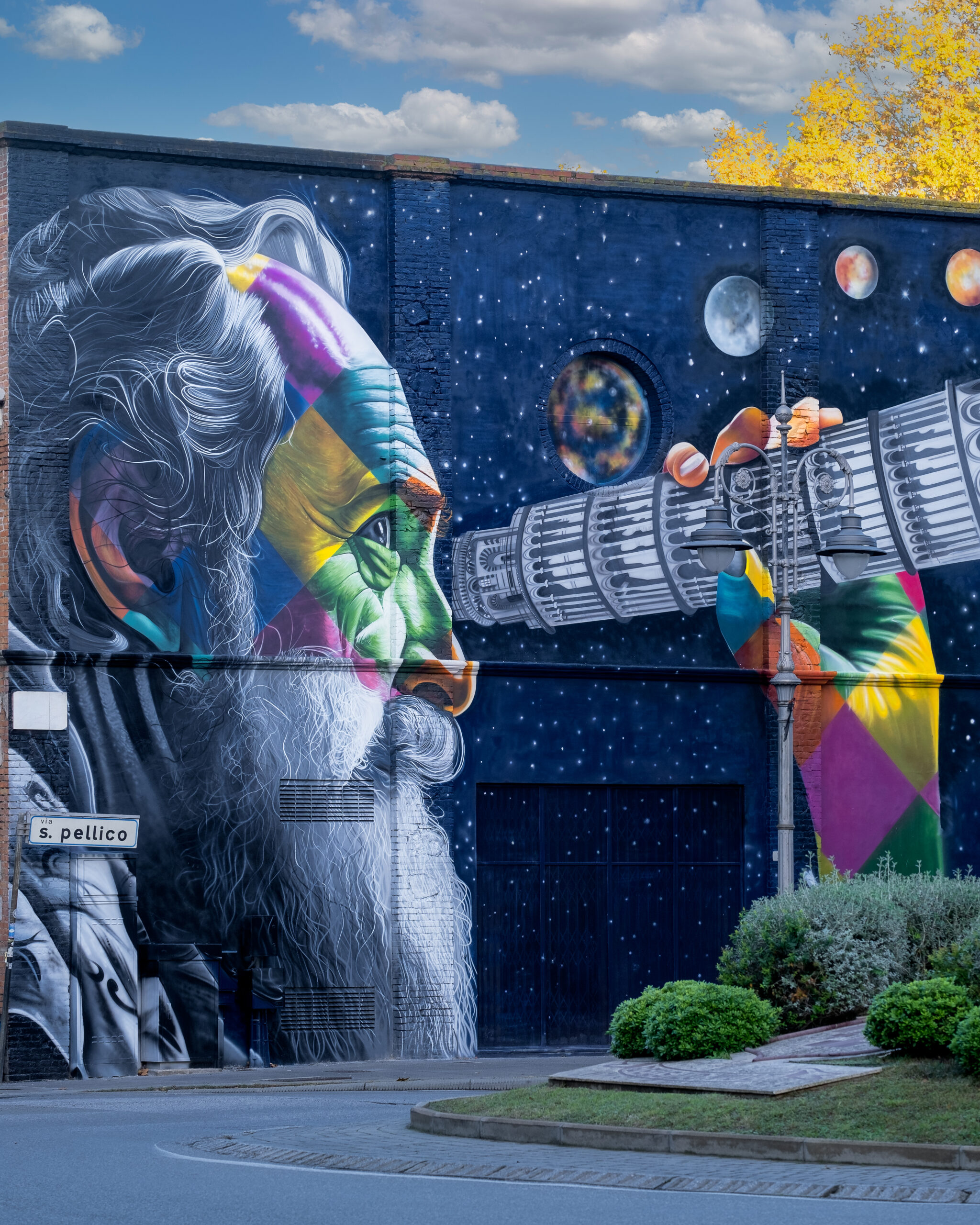 Murale colorato su una facciata urbana raffigurante Galileo Galilei che osserva il cielo con un telescopio, tra stelle, pianeti e dettagli architettonici.