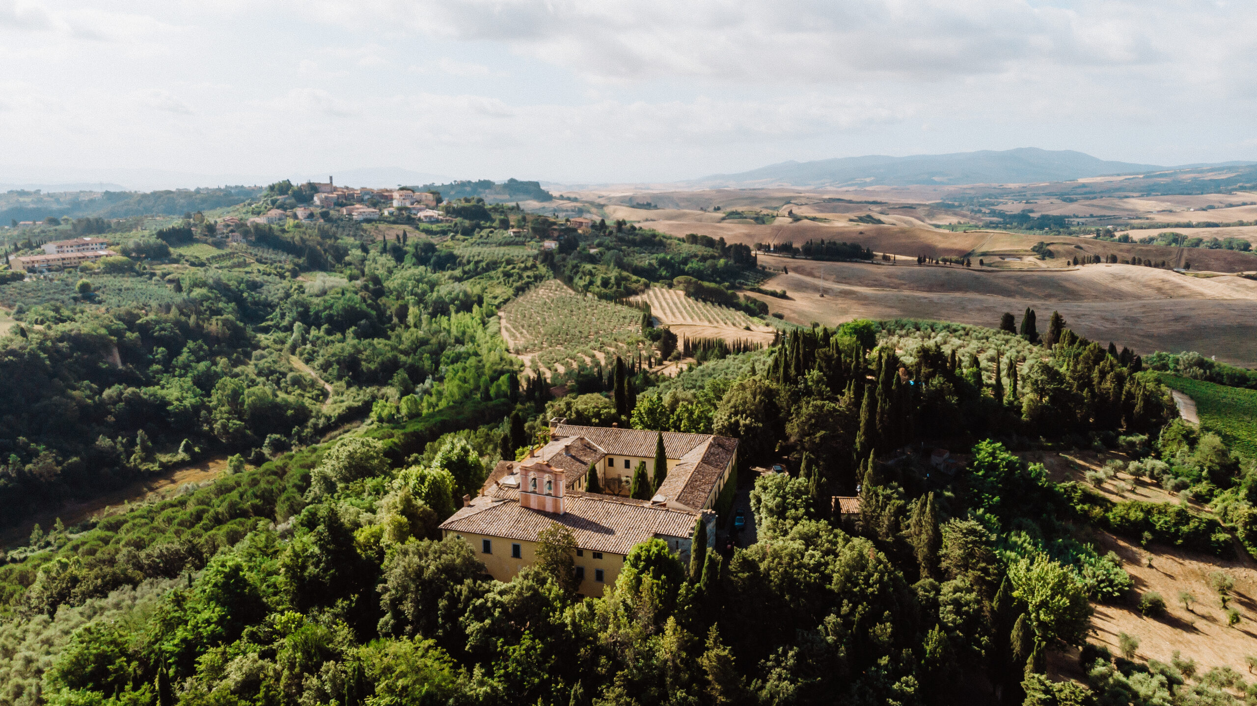 Veduta aerea di Badia di Morrona, immersa nel verde, circondata da cipressi, uliveti e campi coltivati, con un borgo e le colline toscane sullo sfondo.