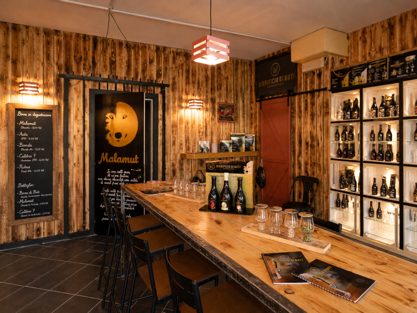 Interno del Birrificio di Buti con pareti in legno, lungo tavolo per le degustazioni, bottiglie di birra artigianale, bicchieri e scaffali espositivi illuminati.