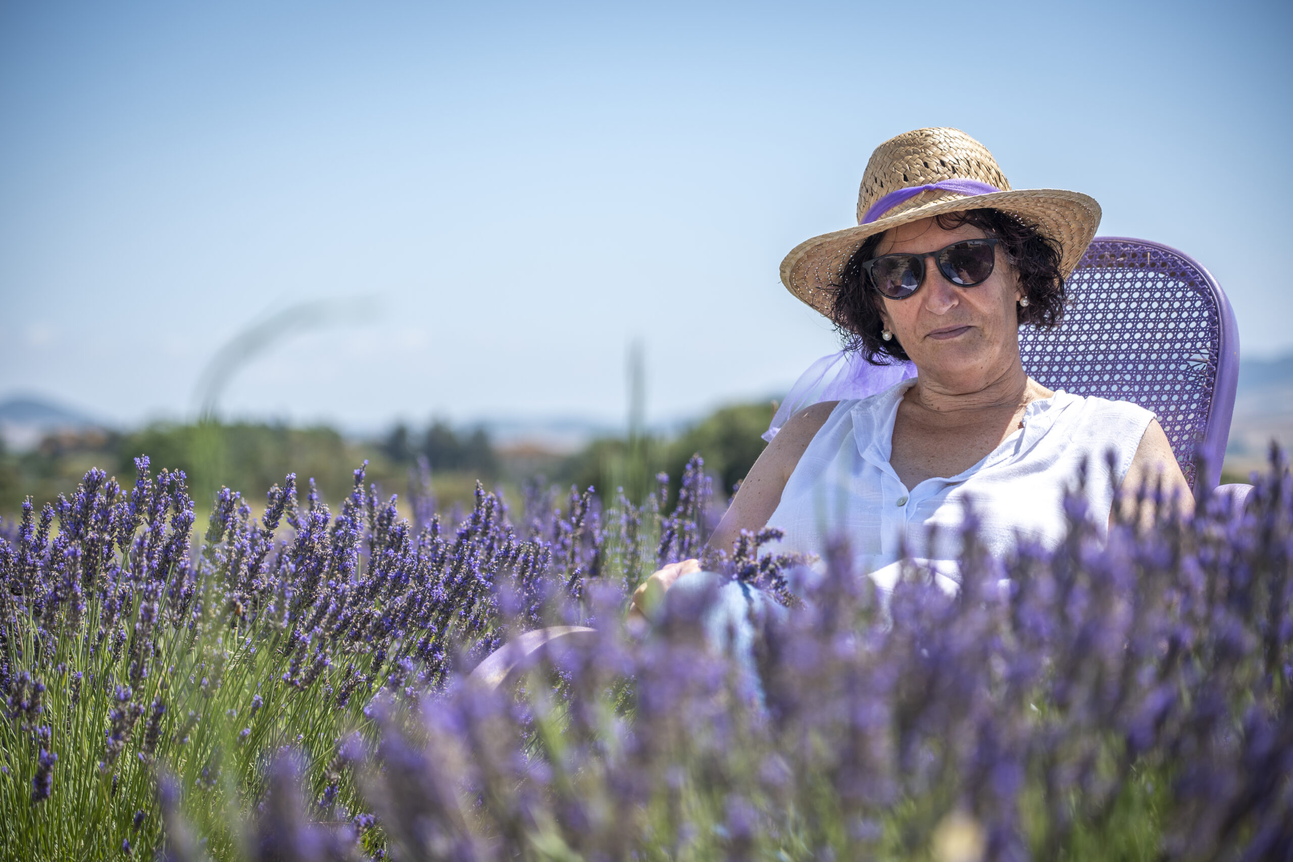 Una donna con cappello di paglia e occhiali da sole siede su una poltrona viola in mezzo a un campo di lavanda, sotto un cielo limpido.