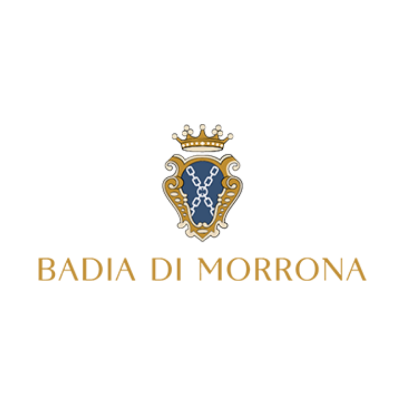 Veduta aerea di Badia di Morrona, immersa nel verde, circondata da cipressi, uliveti e campi coltivati, con un borgo e le colline toscane sullo sfondo.