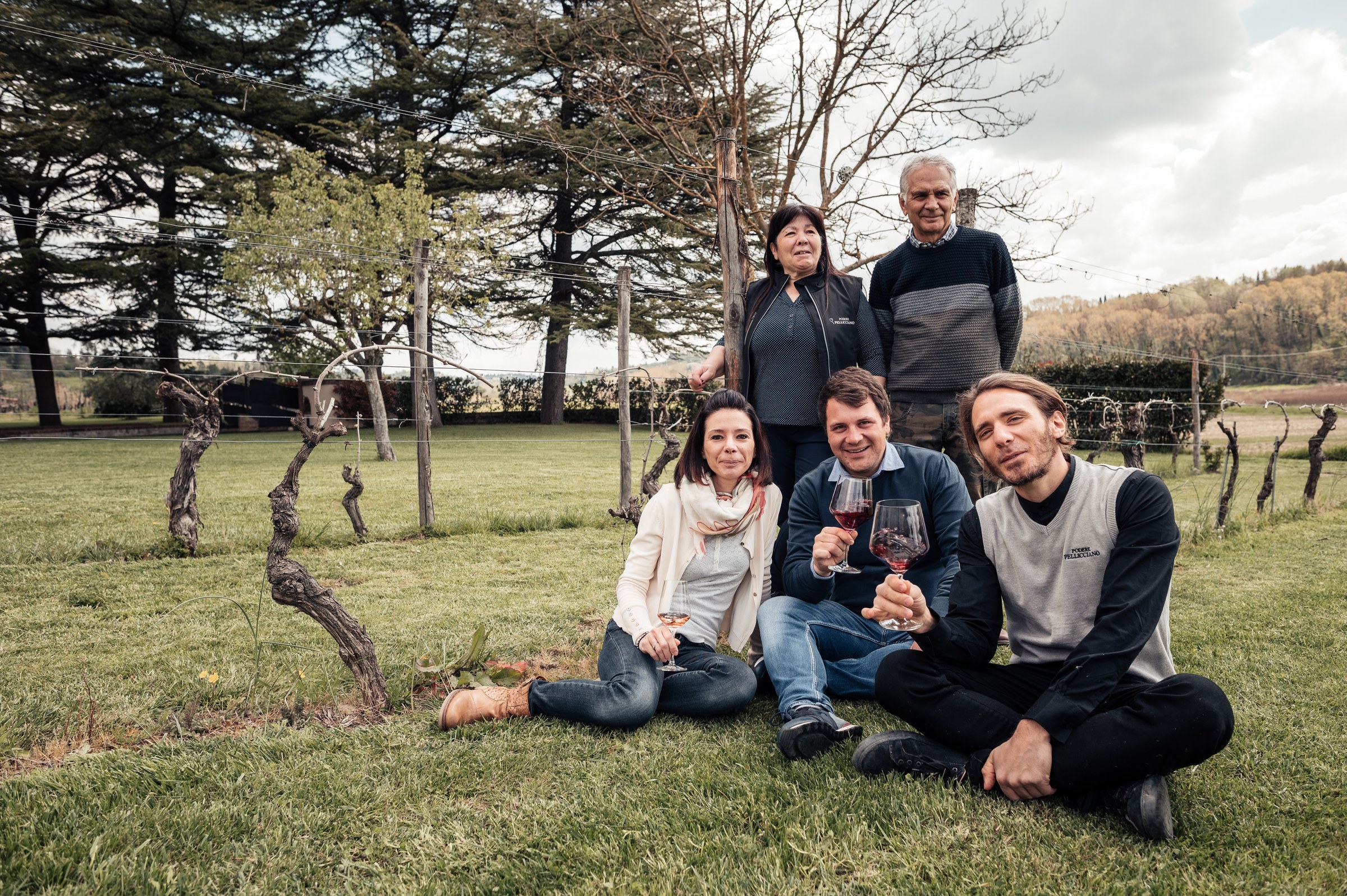 Famiglia di Podere Pellicciano ritratta tra i filari della tenuta sulle colline di San Miniato; alcune persone tengono in mano calici di vino rosso.