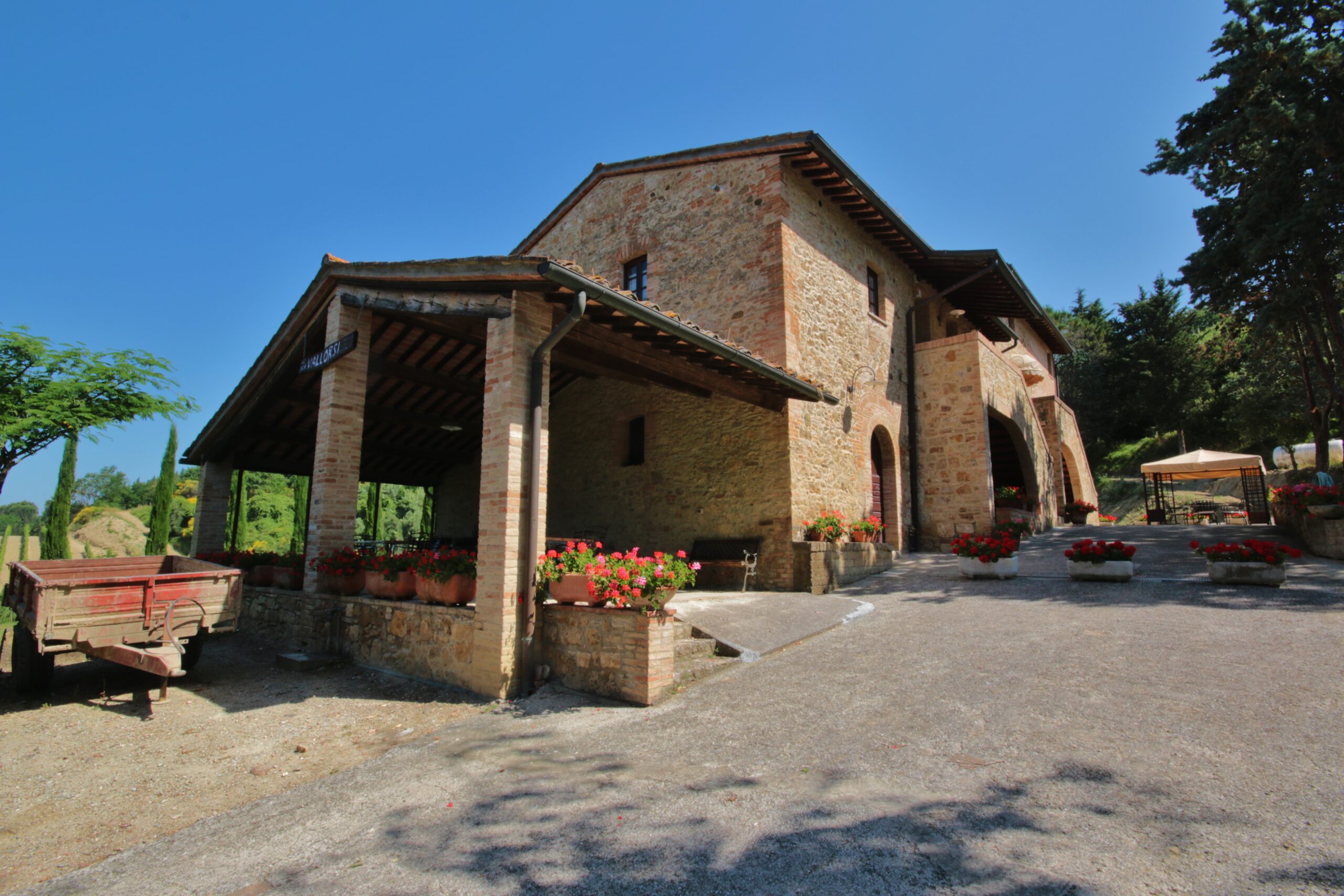 Edificio rurale in pietra dell’Azienda Agricola Vallorsi, con porticato, vasi di fiori rossi e spazi esterni immersi nel verde delle colline pisane.