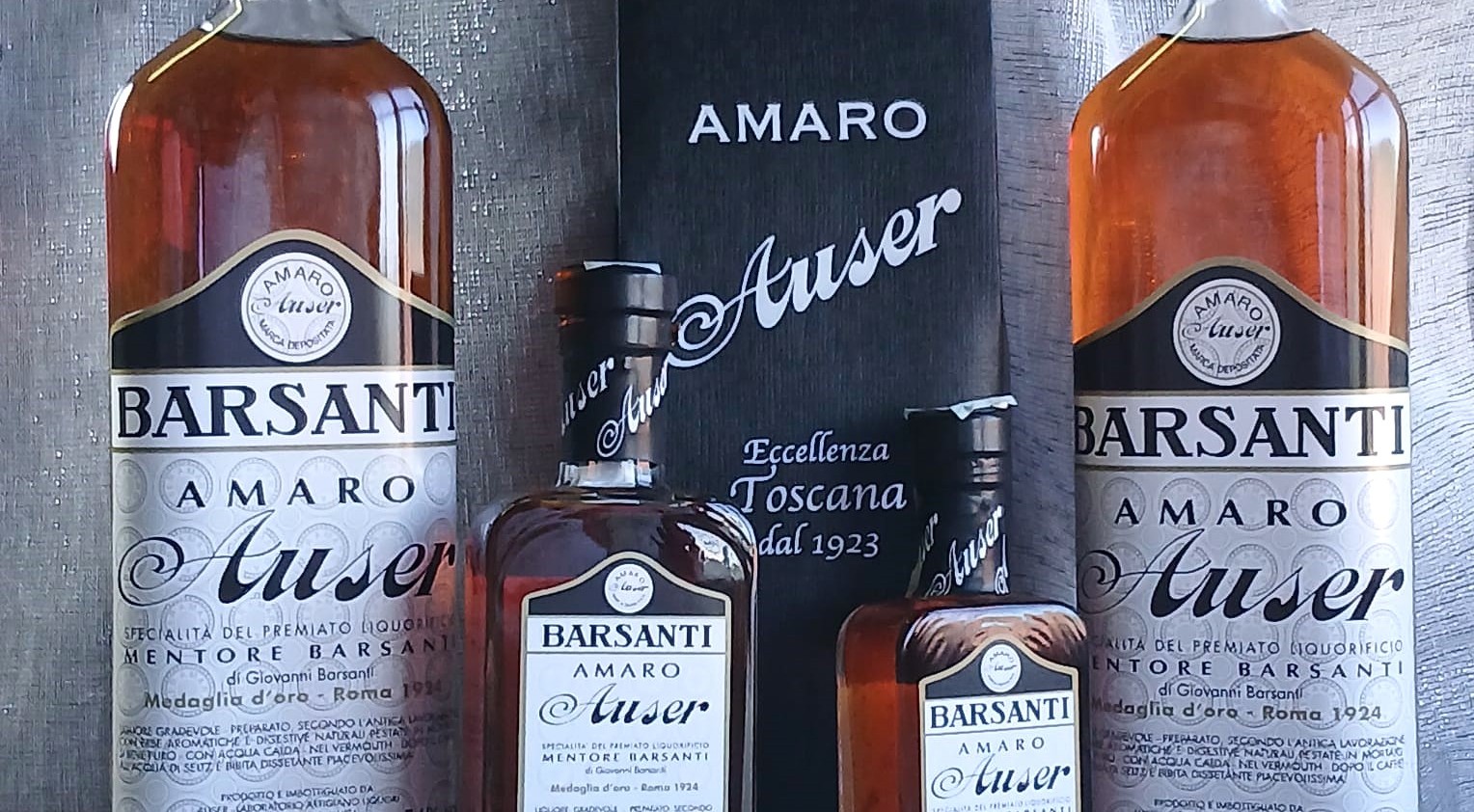 Quattro bottiglie di Amaro Auser Barsanti in diversi formati, affiancate da una confezione nera con la scritta “Eccellenza Toscana dal 1923”.
