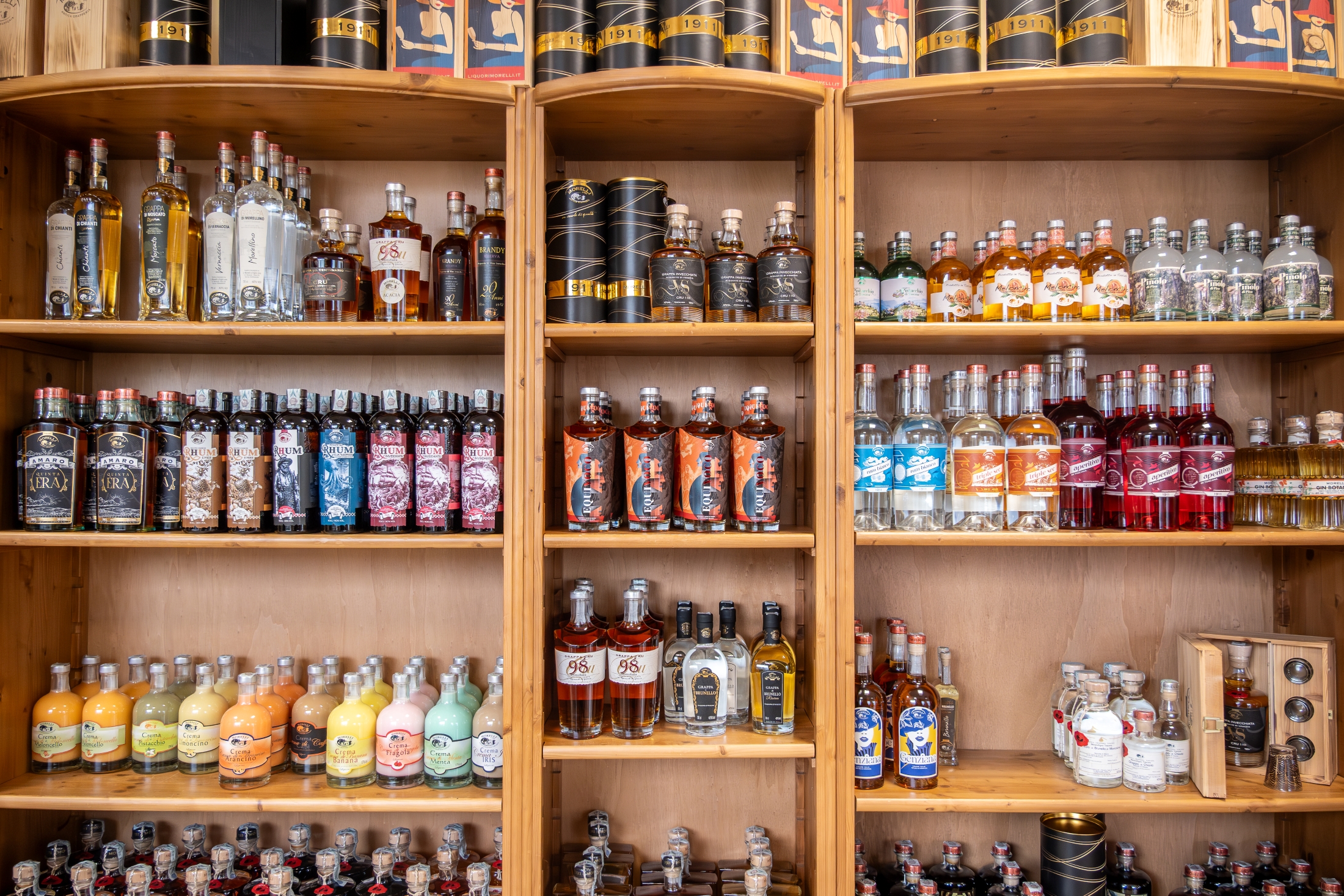 Scaffali in legno con un’ampia selezione di liquori e distillati Liquori Morelli, disposti in bottiglie di diversi formati, colori ed etichette