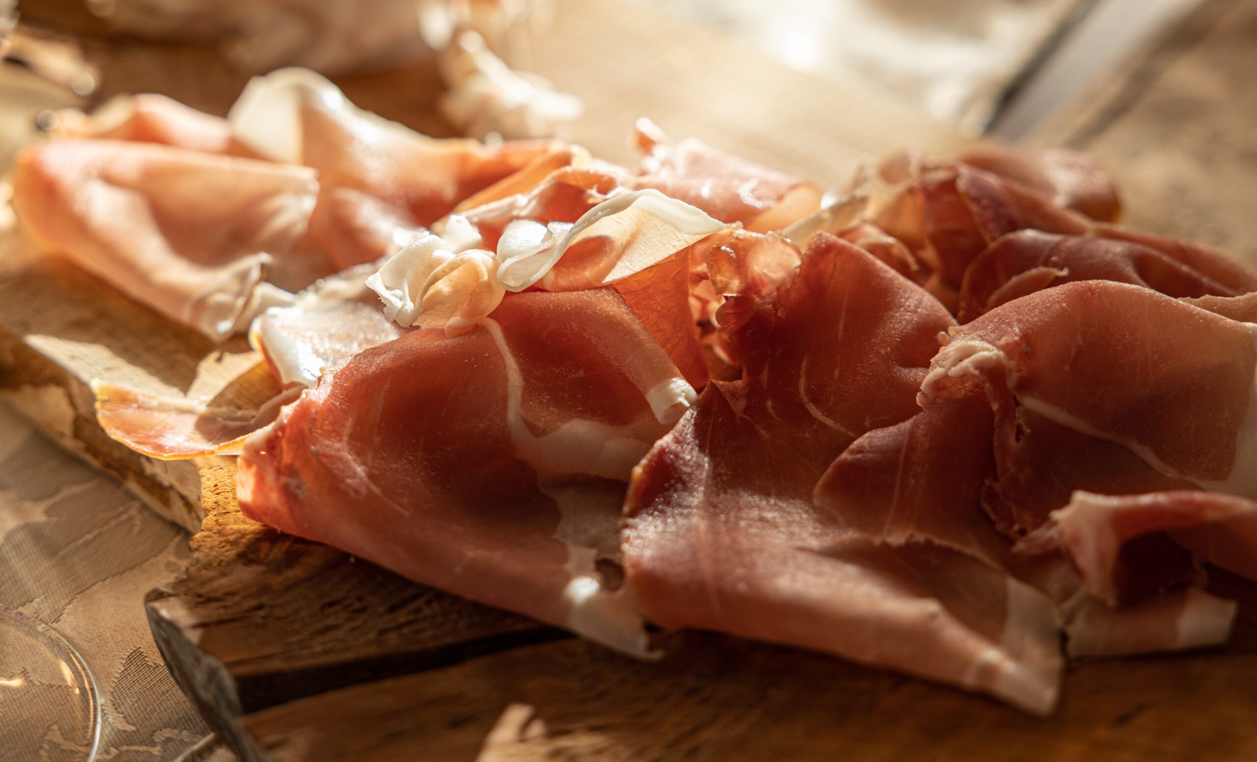 Tagliere di legno con sottili fette di prosciutto crudo e altri salumi, illuminati dalla luce calda del sole su una tavola apparecchiata.