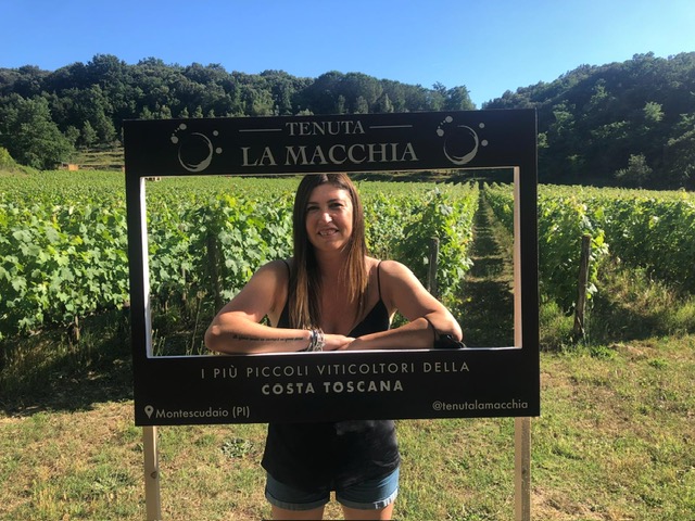 Una donna posa sorridente tra i filari della Tenuta La Macchia, affacciata all’interno di una cornice con il nome dell’azienda e la scritta “I più piccoli viticoltori della Costa Toscana”. Sullo sfondo si vedono il vigneto, la vegetazione e il cielo azzurro.