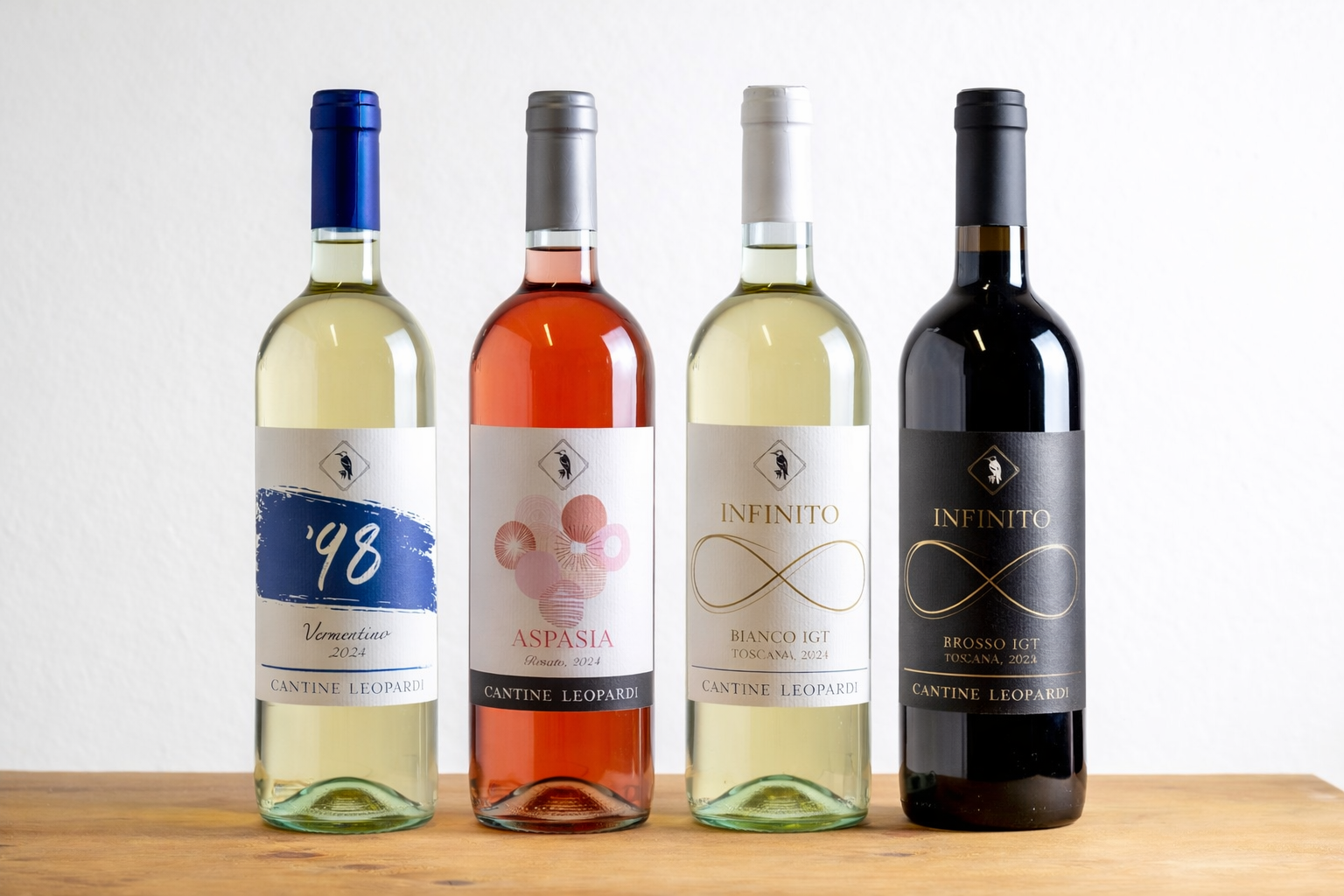 Quattro bottiglie di vino Cantine Leopardi disposte su un piano in legno: Vermentino ’48, Aspasia Rosato, Infinito Bianco IGT Toscana e Infinito Rosso IGT Toscana, su sfondo bianco.