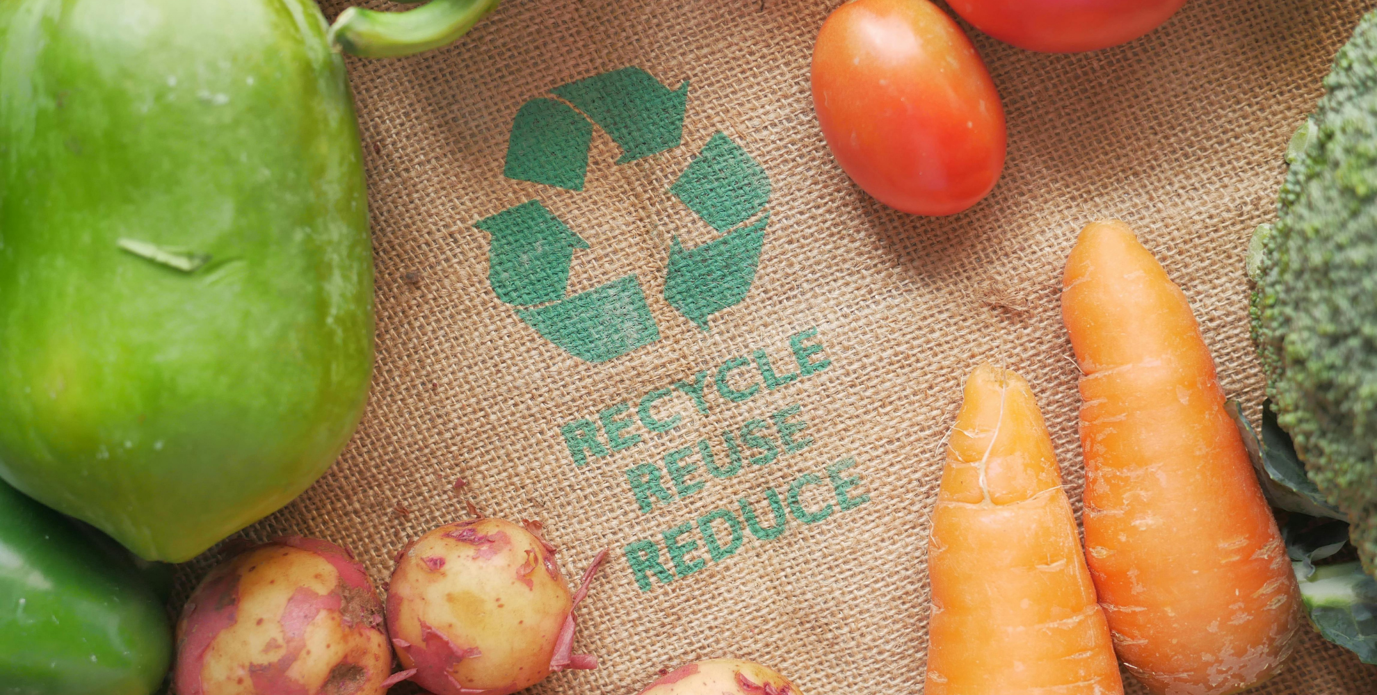 Verdure fresche disposte su un tessuto in juta con simbolo del riciclo e le parole “Recycle, Reuse, Reduce”, a rappresentare sostenibilità, economia circolare e riduzione dei rifiuti.
