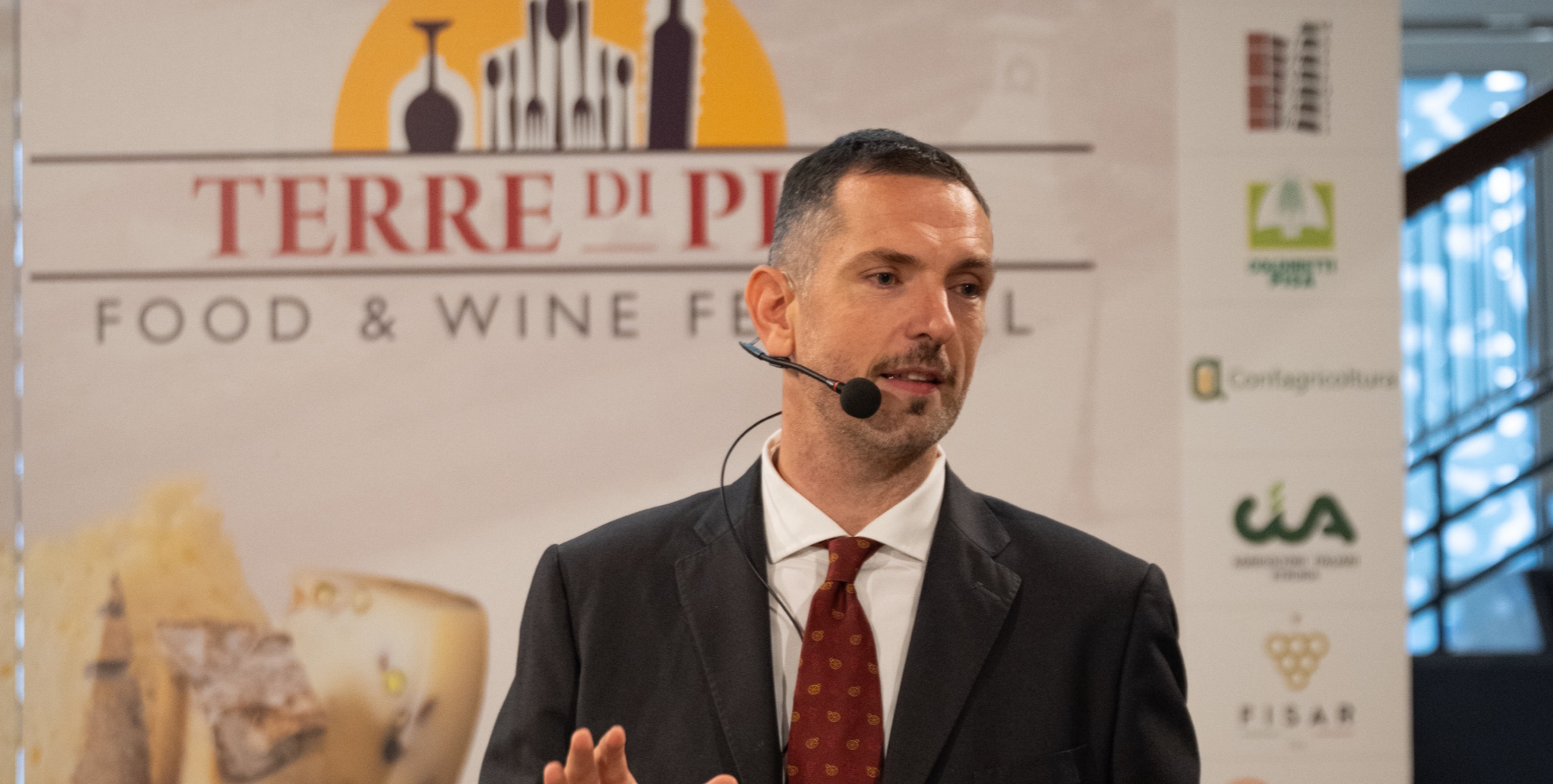 Dario Pantani, in abito scuro con cravatta rossa e microfono ad archetto durante un intervento al Terre di Pisa Food & Wine Festival, davanti al pannello ufficiale dell’evento con i loghi dei partner.