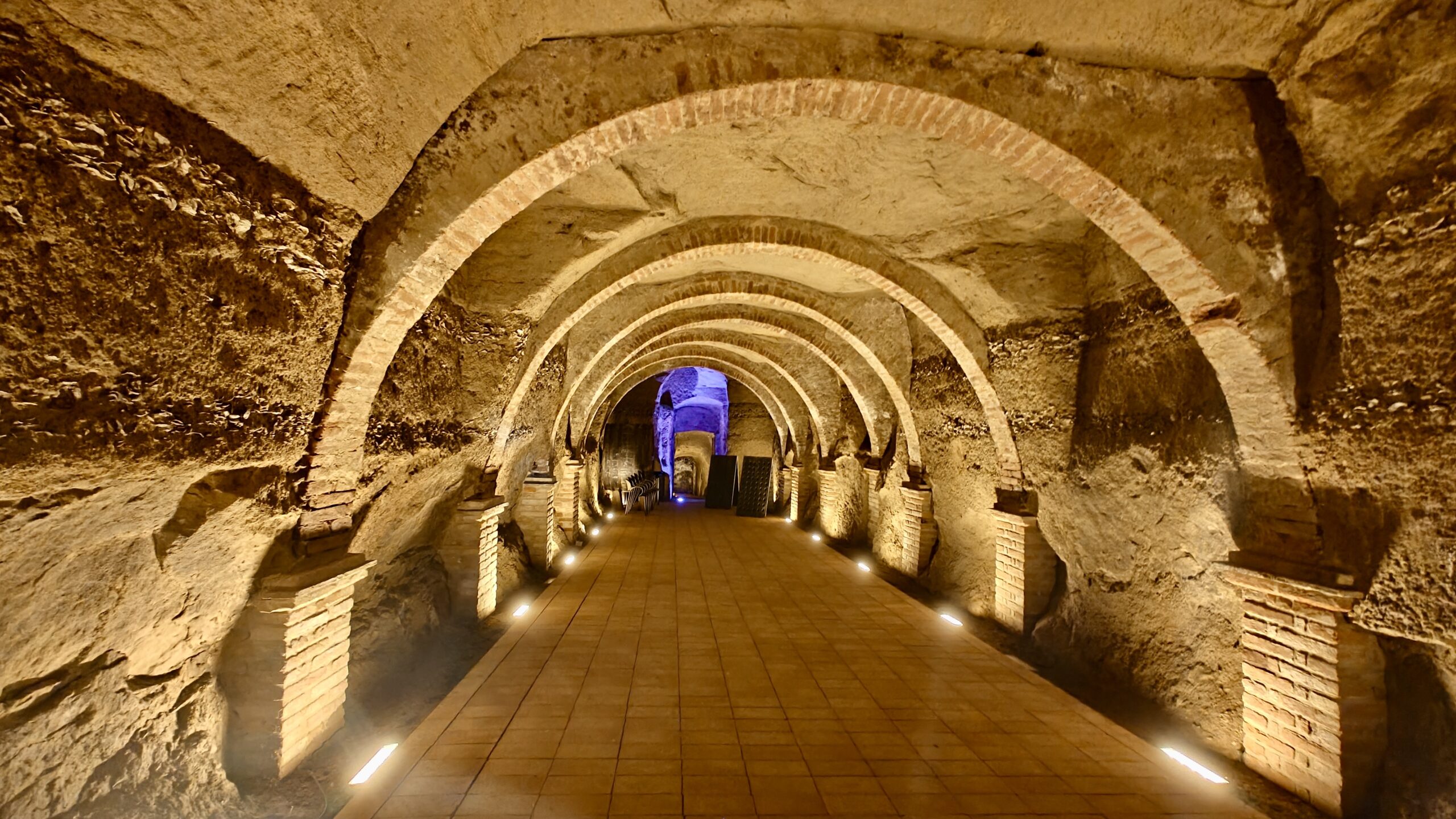 Antica galleria sotterranea in pietra, scandita da una successione di archi in mattoni e illuminata da faretti lungo il pavimento.