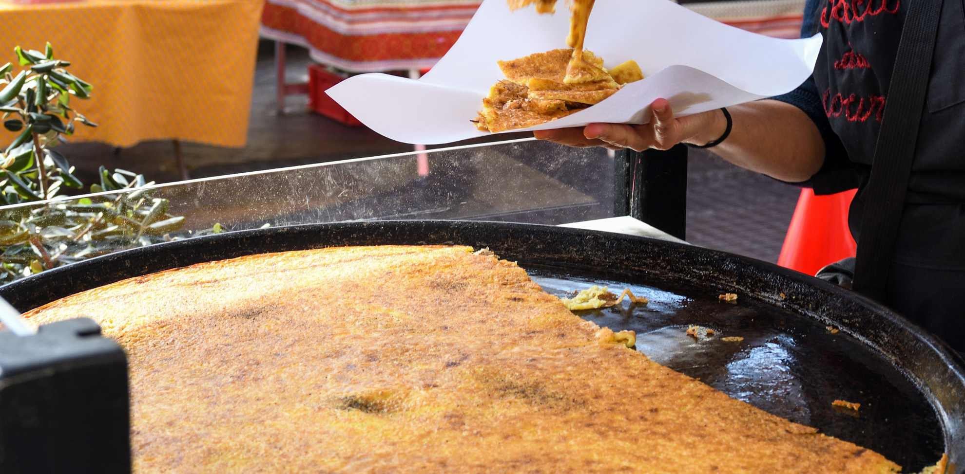 Una persona serve una porzione di cecina appena cotta, tagliata a pezzi e disposta su un foglio di carta, accanto alla grande teglia usata per la cottura.