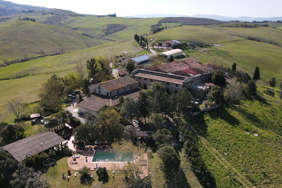 Veduta aerea della Fattoria Lischeto, immersa tra le colline toscane, con gli edifici rurali, la piscina e i filari di cipressi.