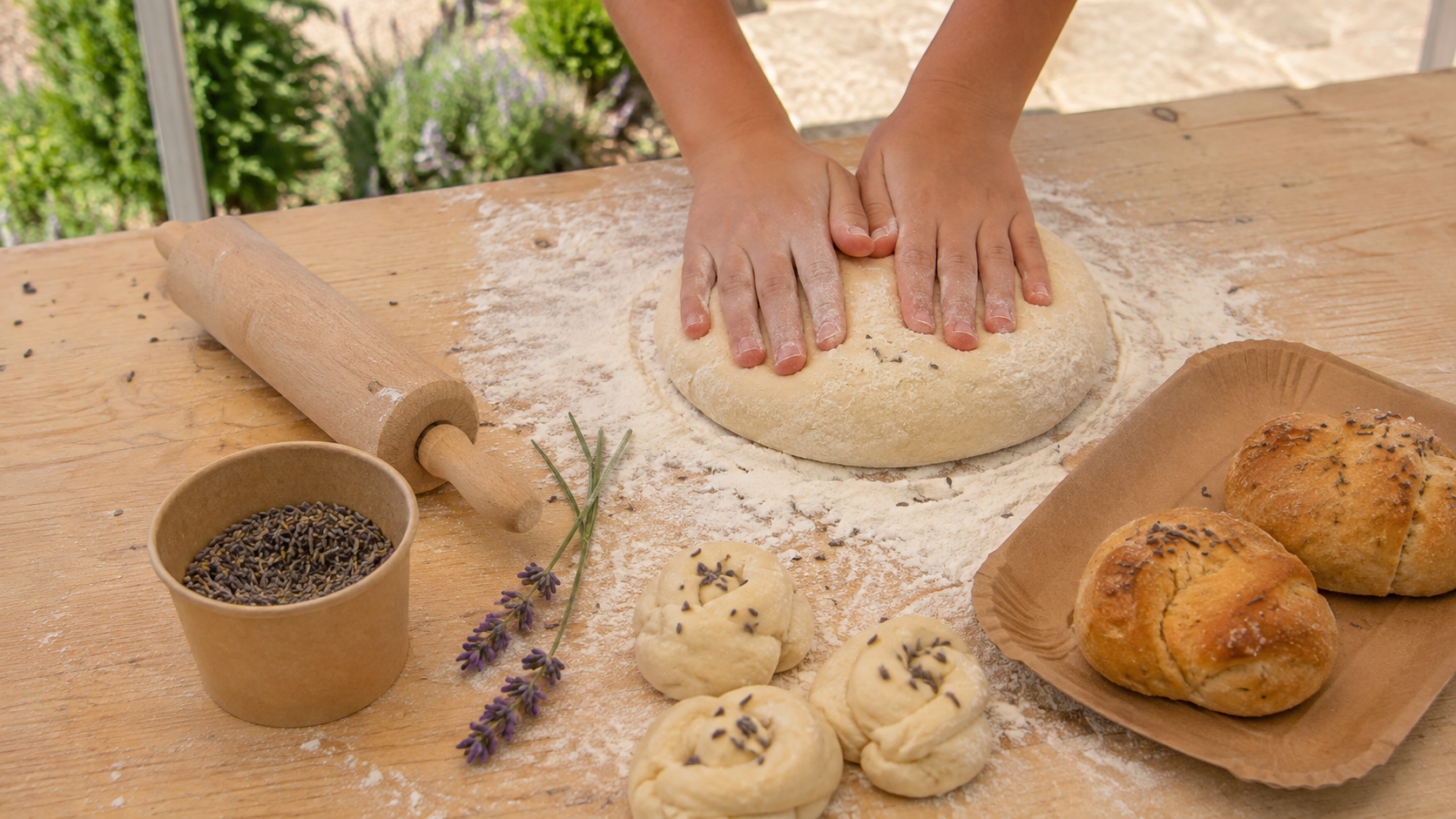 Mani di un bambino lavorano un impasto su un tavolo infarinato all’aperto, accanto a lavanda alimentare, piccoli panini crudi e panificati appena cotti.