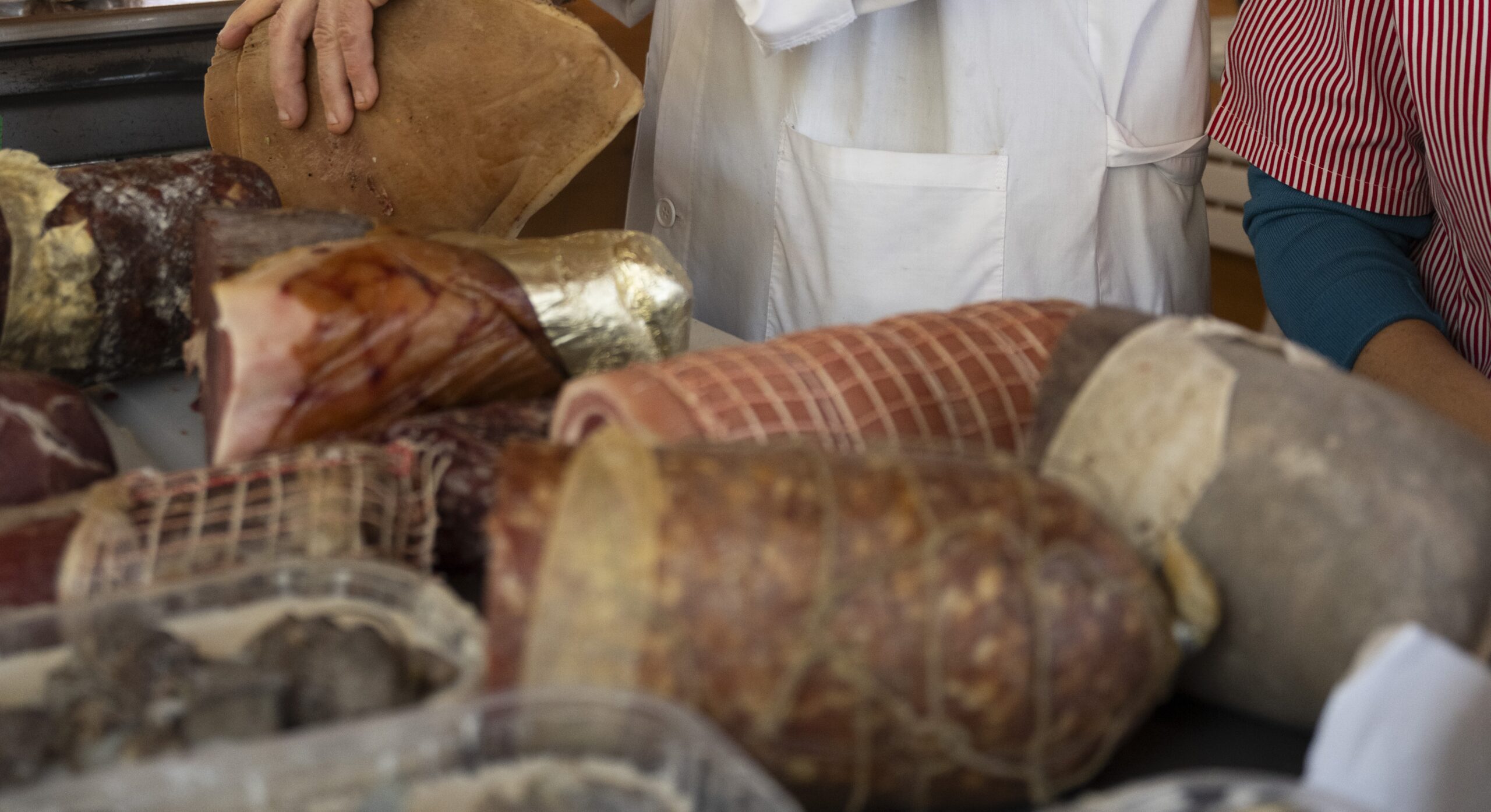 Banco di macelleria con salumi e insaccati artigianali, mentre due addetti in camice bianco servono i prodotti.