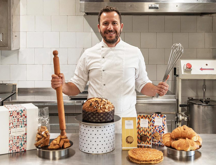 Pasticcere nel laboratorio di DolceMania Italia, circondato da panettone, biscotti, croissant e altri prodotti di pasticceria artigianale.