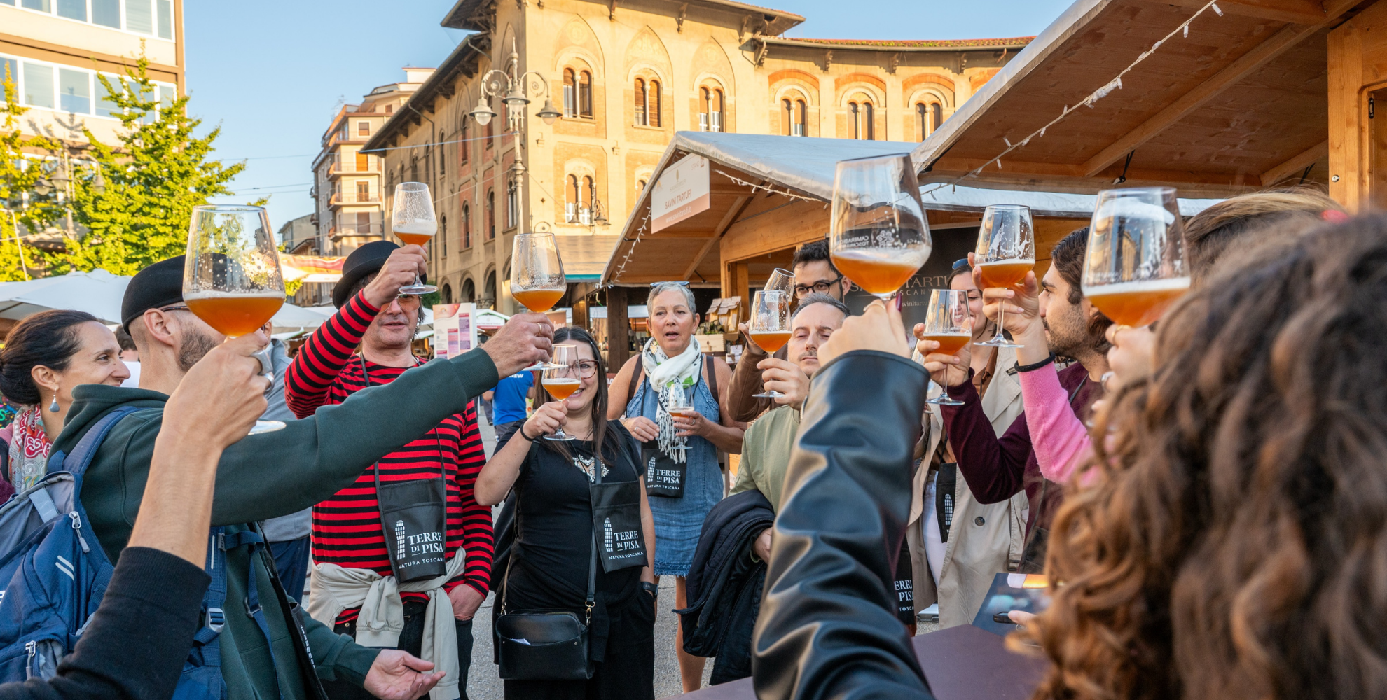 Gruppo di partecipanti durante una degustazione di birra all’aperto al Terre di Pisa Food & Wine Festival, tra gli stand di Piazza Vittorio Emanuele II a Pisa, con calici di birra sollevati per un brindisi.