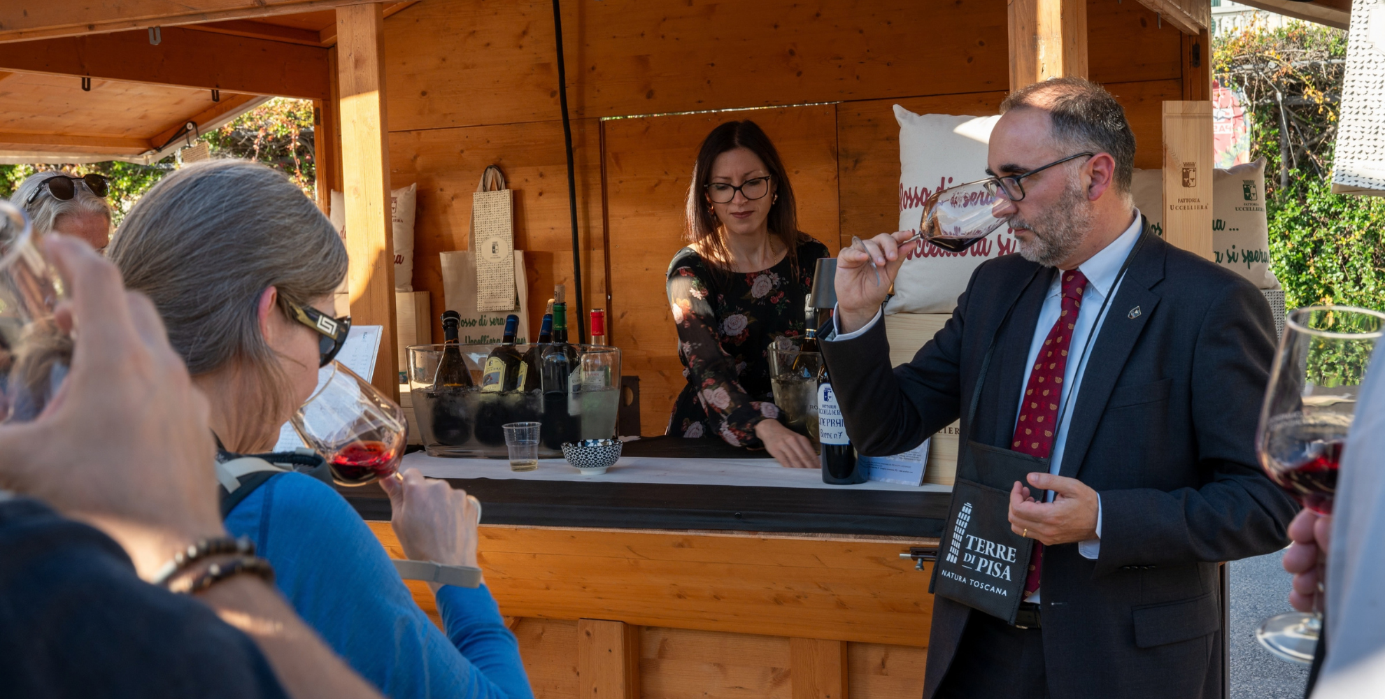 Sommelier guida una degustazione di vino davanti allo stand di una cantina durante il Terre di Pisa Food & Wine Festival, mentre i partecipanti osservano e assaggiano i vini proposti.