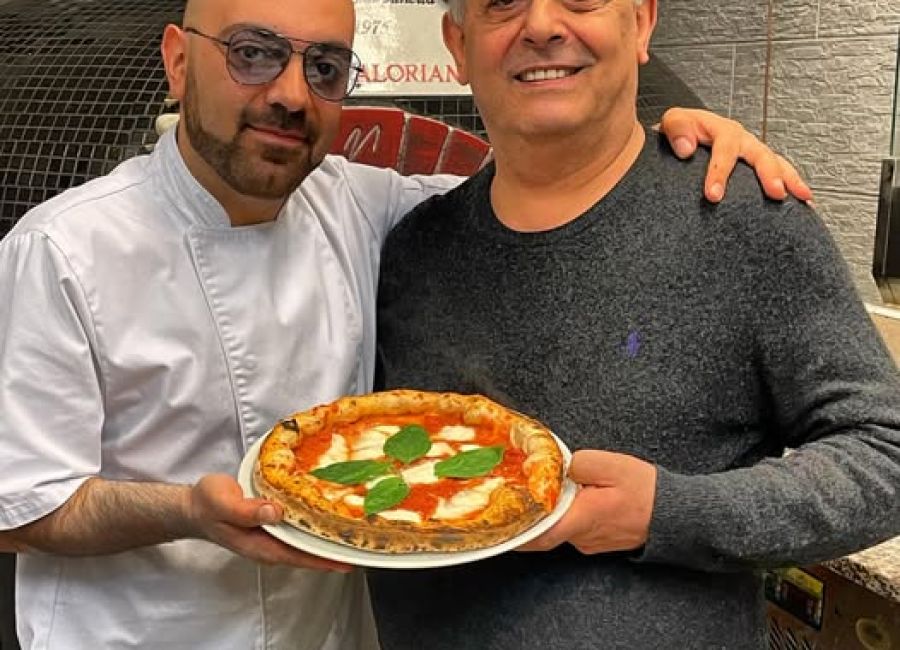 Un pizzaiolo in divisa bianca e un uomo sorridente mostrano una pizza margherita appena sfornata, all’interno di una pizzeria.