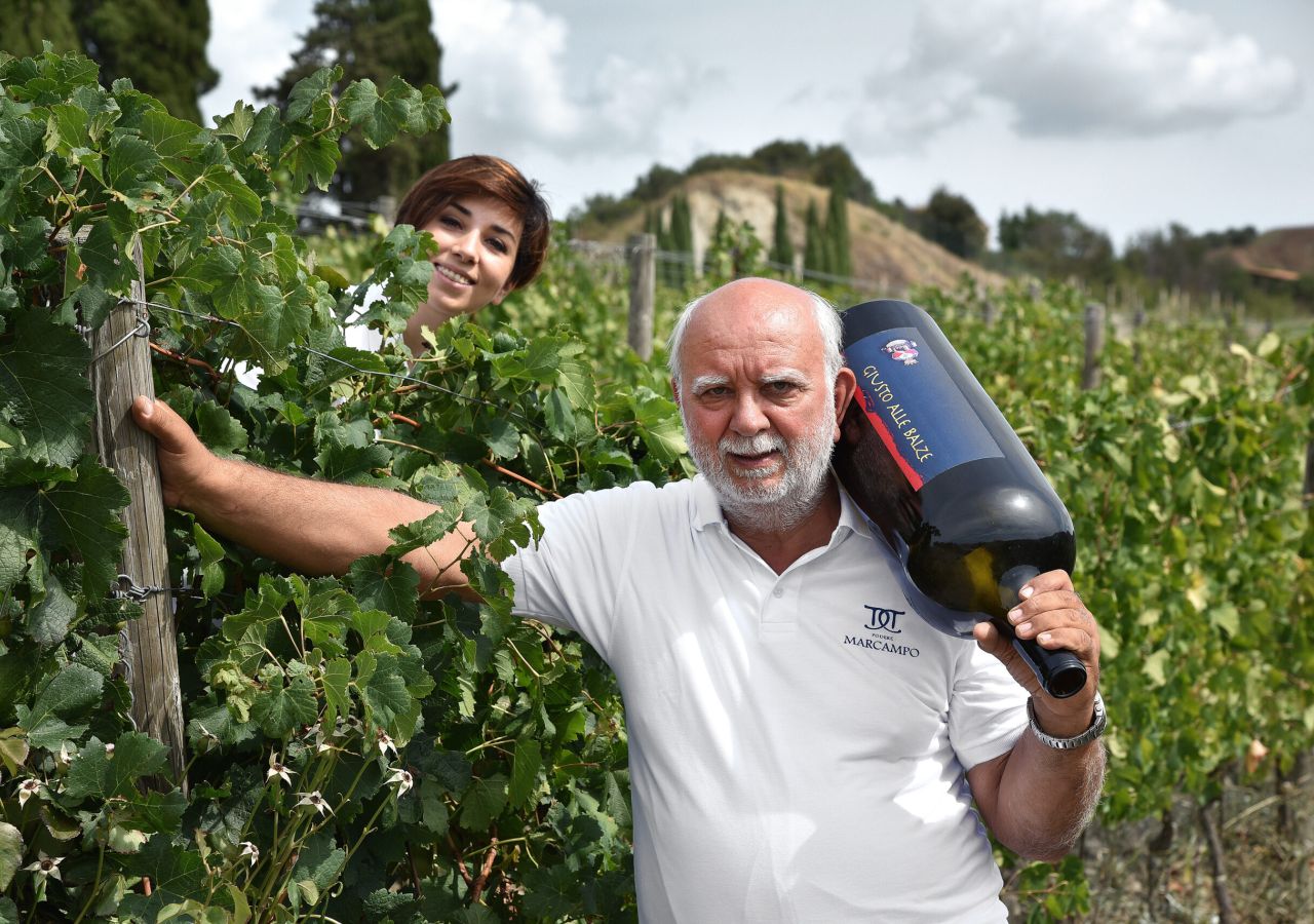 Un uomo con una grande bottiglia di vino sulla spalla e una donna tra i filari posano sorridenti nel vigneto di Podere Marcampo.