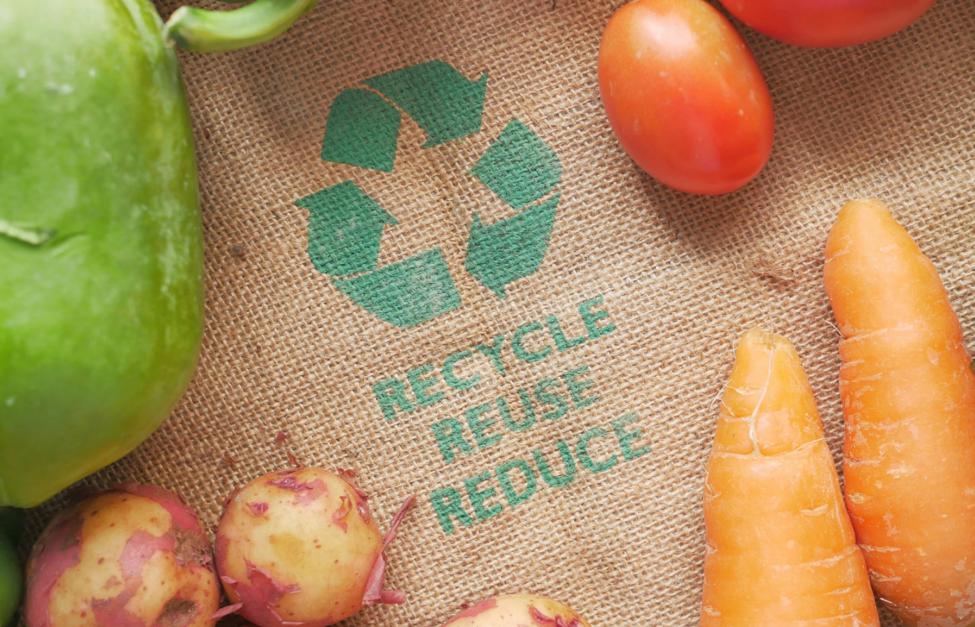 Verdure fresche disposte su un tessuto in juta con simbolo del riciclo e le parole “Recycle, Reuse, Reduce”, a rappresentare sostenibilità, economia circolare e riduzione dei rifiuti.