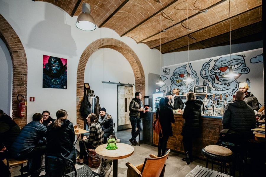 Interno della Tap Room del Birrificio La Staffetta a Calci, con clienti ai tavoli e al bancone sotto le volte in mattoncini.
