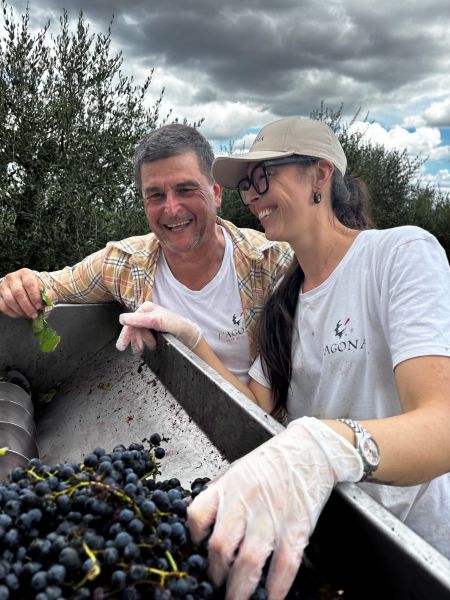 Donna sorridente tra i filari della tenuta L’Agona mentre solleva un calice di vino bianco.