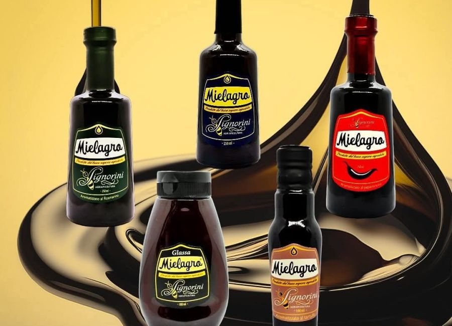 Cinque confezioni di Mielagro dell’Agriapicoltura Signorini, in diverse varianti e formati, disposte su uno sfondo dorato con una colata scura e lucida.