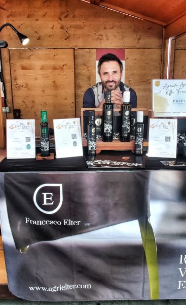 Un uomo dietro lo stand dell’Azienda Agricola Elter, con bottiglie di olio extravergine di oliva, confezioni e materiali informativi esposti sul banco.