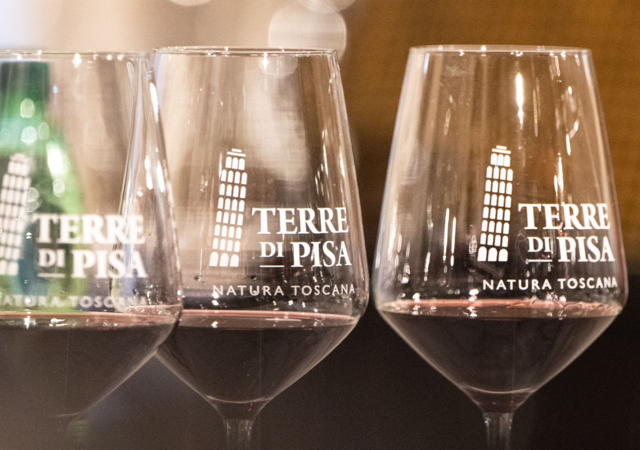 Calici da degustazione con il logo Terre di Pisa, alcuni contenenti vino rosso, disposti in primo piano durante una masterclass enologica.