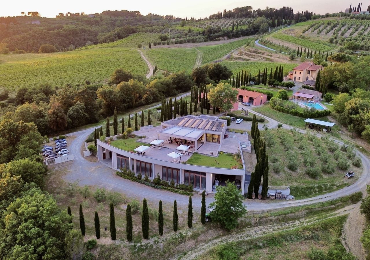 Veduta aerea della Tenuta Le Palaie, immersa nelle colline di Peccioli, tra vigneti, oliveti e filari di cipressi, con la cantina, l’agriturismo e la piscina circondati dal verde.