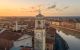 Veduta aerea di Pisa al tramonto, con la torre dell’orologio di Palazzo Pretorio in primo piano e i Lungarni lungo il fiume Arno sullo sfondo.
