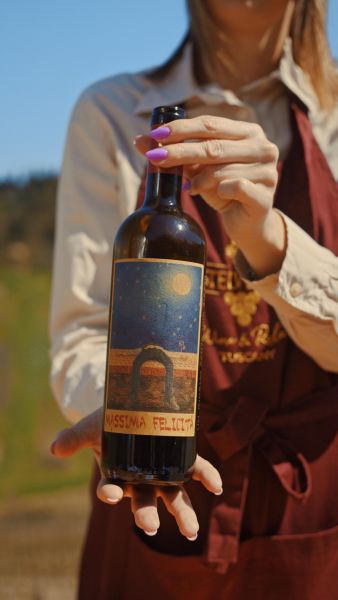 Quattro donne con grembiuli color bordeaux camminano tra i filari al tramonto, portando cassette e bottiglie di vino, in un paesaggio collinare toscano con cipressi e oleandri in fiore.