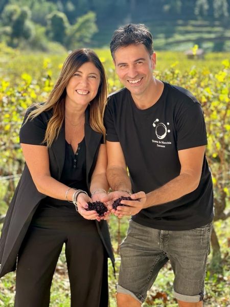 Una donna e un uomo sorridono tra i filari di un vigneto, mostrando grappoli d’uva scura raccolti nelle mani. L’uomo indossa una maglietta con il logo di Tenuta La Macchia.