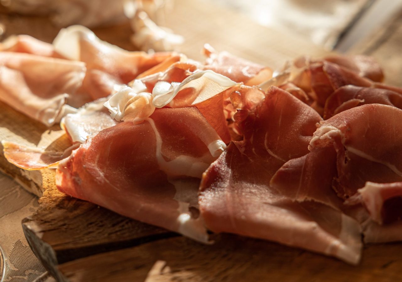 Tagliere di legno con sottili fette di prosciutto crudo e altri salumi, illuminati dalla luce calda del sole su una tavola apparecchiata.