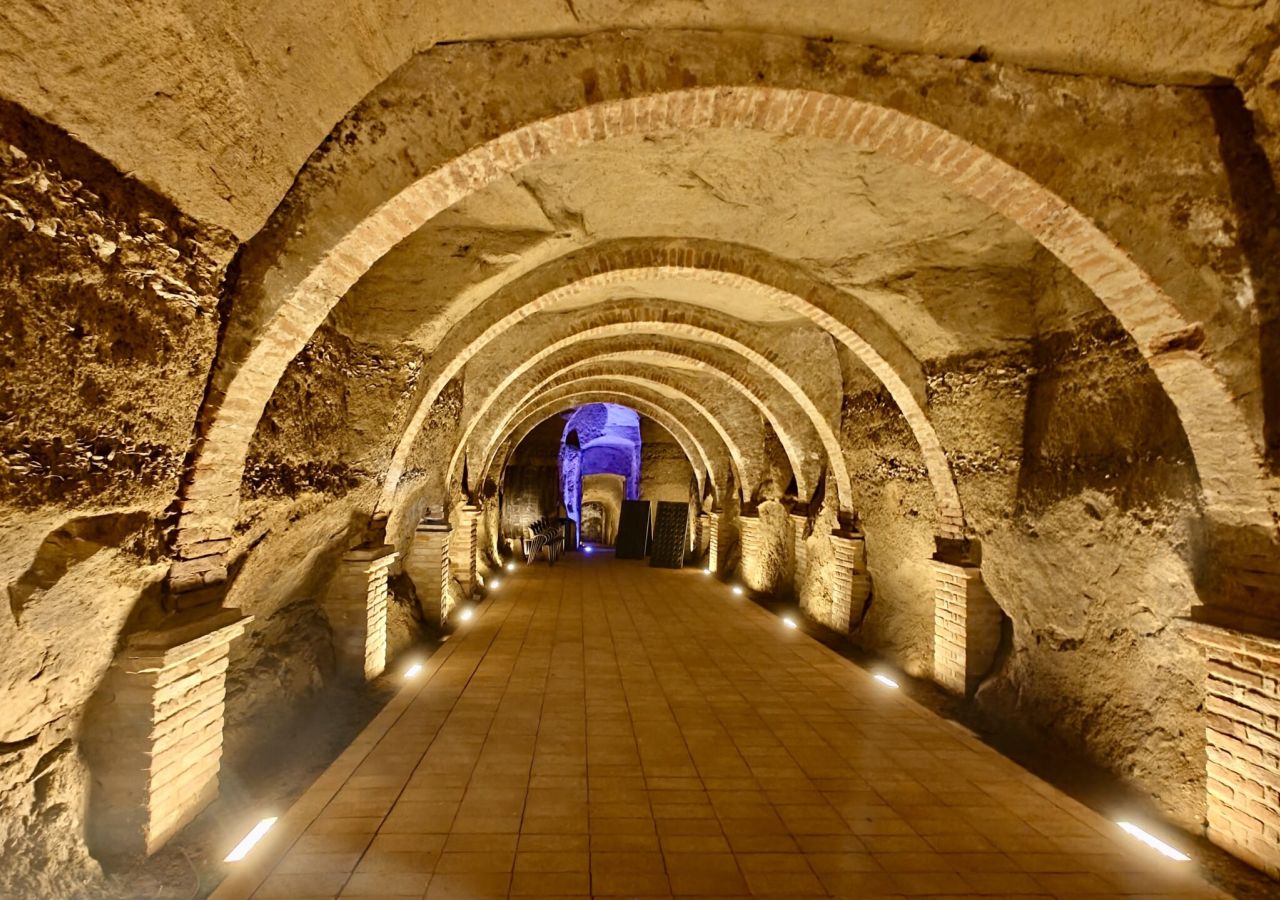Antica galleria sotterranea in pietra, scandita da una successione di archi in mattoni e illuminata da faretti lungo il pavimento.