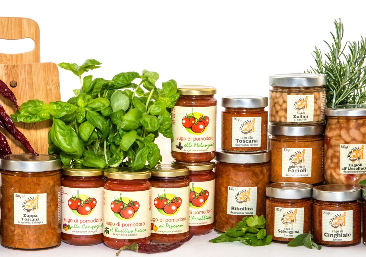 Assortimento di conserve e specialità toscane in barattolo di vetro, tra cui sughi di pomodoro, ragù, zuppe, legumi e salse per crostini, disposti con basilico, rosmarino, salvia e peperoncini secchi.