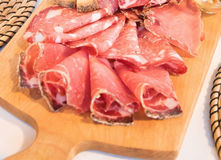 Tagliere di legno con assortimento di salumi affettati, formaggi stagionati e una ciotolina di miele con pinoli.