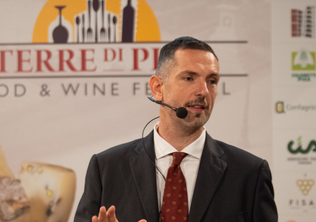 Dario Pantani, in abito scuro con cravatta rossa e microfono ad archetto durante un intervento al Terre di Pisa Food & Wine Festival, davanti al pannello ufficiale dell’evento con i loghi dei partner.