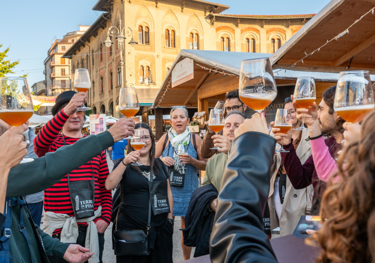 Gruppo di partecipanti durante una degustazione di birra all’aperto al Terre di Pisa Food & Wine Festival, tra gli stand di Piazza Vittorio Emanuele II a Pisa, con calici di birra sollevati per un brindisi.
