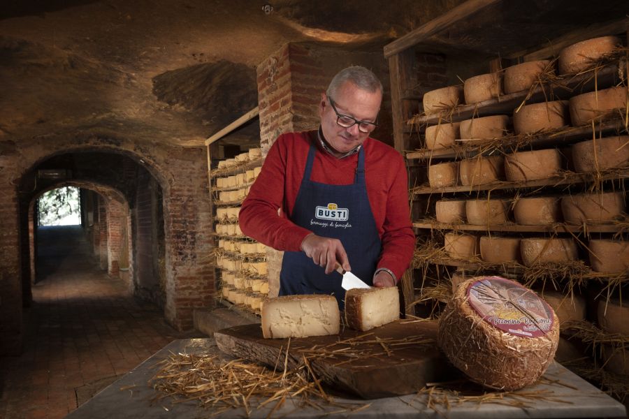 Un uomo con grembiule del Caseificio Busti taglia una forma di Pecorino Roncione in una cantina di stagionatura, circondato da scaffali colmi di formaggi affinati nella paglia.