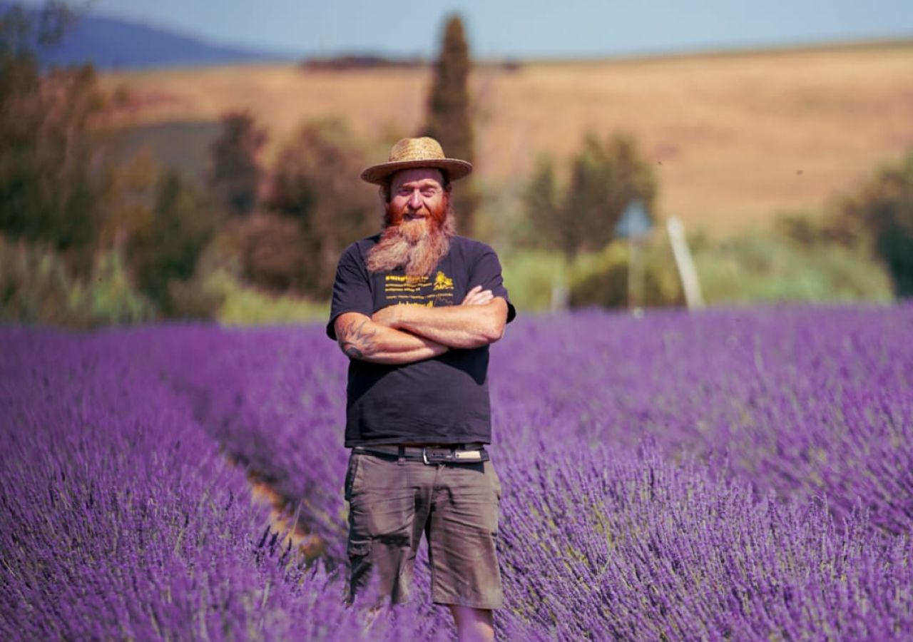 Uomo con barba e cappello di paglia in piedi tra i filari di lavanda in fiore, con le colline toscane sullo sfondo.