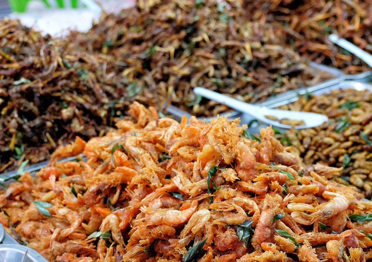 Banco gastronomico con grandi vassoi metallici pieni di alimenti fritti disposti in mucchi. In primo piano si vede un vassoio colmo di piccoli crostacei fritti, mentre sullo sfondo compaiono altri vassoi con insetti e larve commestibili, alcuni con pinze da servizio appoggiate sopra. La scena è ripresa da vicino e trasmette l’idea di un’esposizione di street food o mercato alimentare.